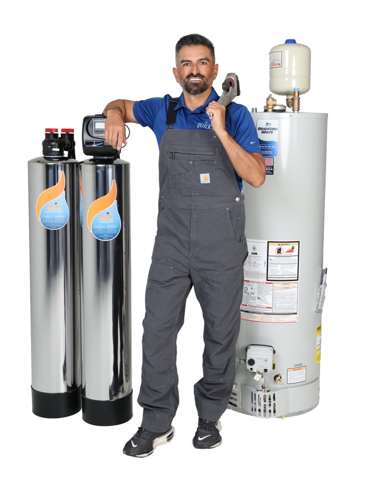 A Quick Water Heater technician in work overalls with a wrench on his shoulder, beside a water heater and filtration tanks